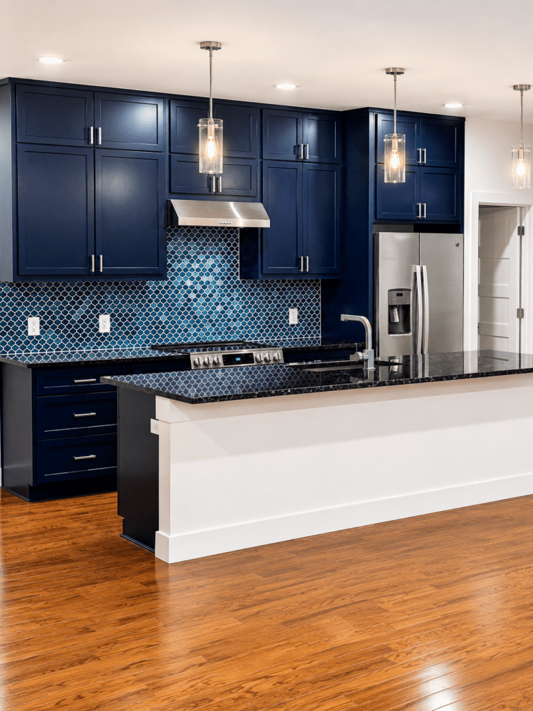 Driftwood kitchen remodel with dark blue cabinets, patterned tile backsplash, black countertops, stainless steel appliances, pendant lighting, and hardwood flooring